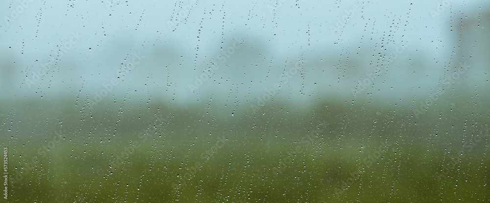 Atmospheric minimal backdrop with rain droplets on glass. Wet window with rainy drops and dirt spots closeup. Blurry buildings and green trees under blue sky against dirty window glass with raindrops. Atmospheric minimal backdrop with rain droplets on glass. Wet window with rainy drops and dirt spots closeup. Blurry buildings and green trees under blue sky against dirty window glass with raindrops.