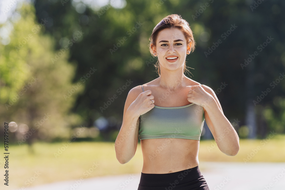 Cheerful caucasian girl in sporty top, workout outdoors brad smiles. Young hispanic brunette girl jogging at park looks at camera. Mockup, fitness, health care. Pretty lady trains on summer morning Cheerful caucasian girl in sporty top, workout outdoors brad smiles. Young hispanic brunette girl jogging at park looks at camera. Mockup, fitness, health care. Pretty lady trains on summer morning