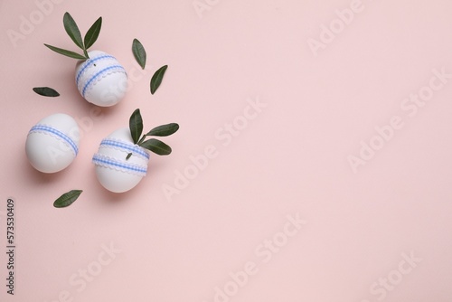 Beautifully decorated Easter eggs and green leaves on pale pink background, flat lay. Space for text