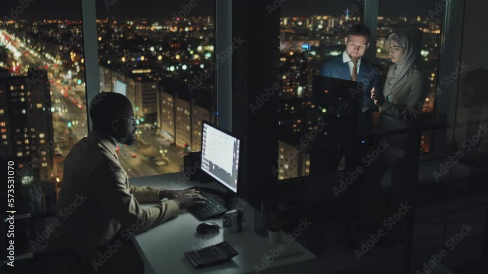Full shot of three employees working overtime at night in city office, Afro-American male typing email on computer, Caucasian man and Muslim woman holding laptop and talking, with streetlights behind Full shot of three employees working overtime at night in city office, Afro-American male typing email on computer, Caucasian man and Muslim woman holding laptop and talking, with streetlights behind