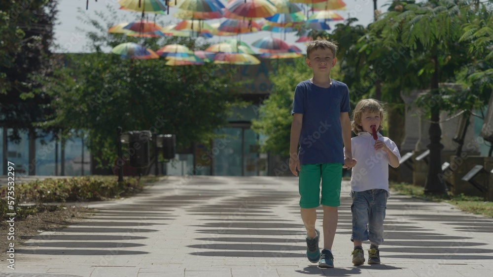 Two brother walking hand in hand under colored suspended umbrella, eat ice cream Two brother walking hand in hand under colored suspended umbrella, eat ice cream