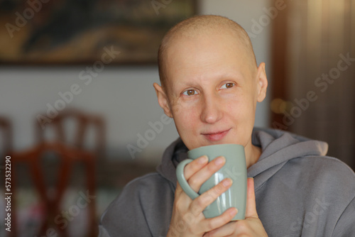 Hairless happy woman is holding cup of hot drink, coffee or tea. Bald female is having remission after cancer disease enjoying breakfast at living room. Pensive relaxed girl is looking in side at home photo