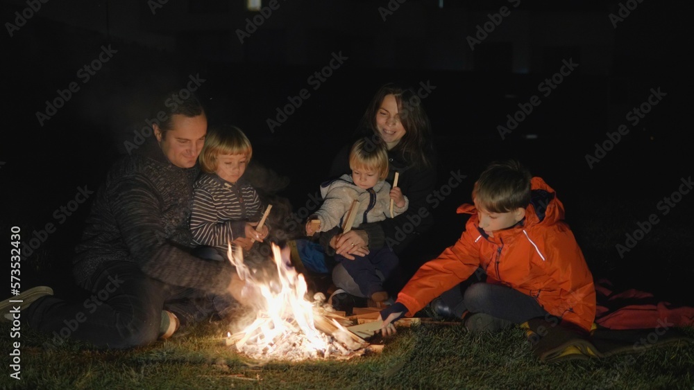 Happy parents and children have fun at campfire, put wood on the fire