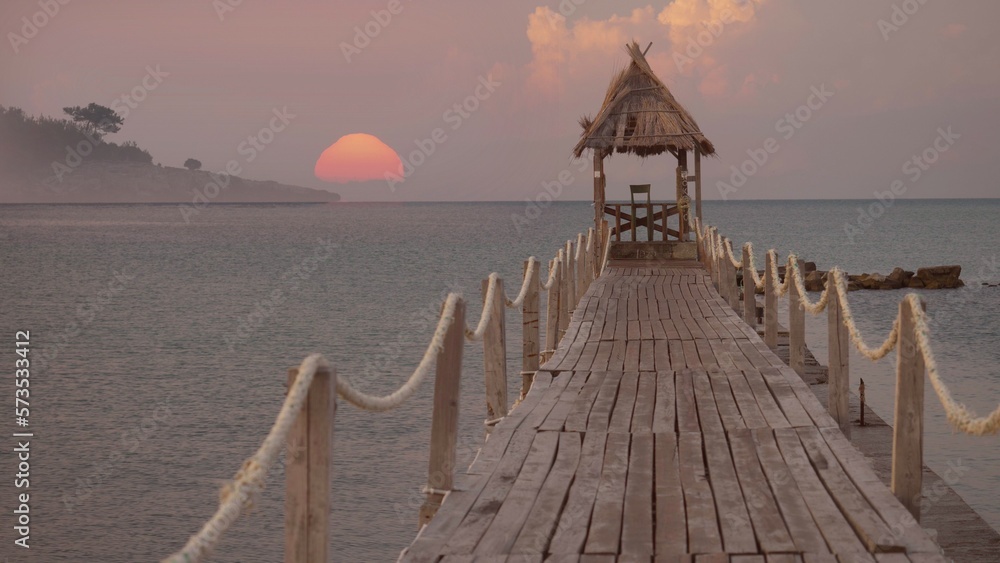 Fototapeta premium Wooden pontoon with end hut into the sea at sunrise over island