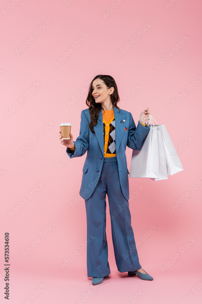 full length of happy woman in blue pantsuit holding shopping bags and coffee to go on pink. full length of happy woman in blue pantsuit holding shopping bags and coffee to go on pink.