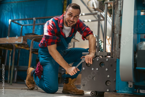 Young male mechanic kneeling on his leg with a smile on his face, while screwing together parts of the metal elements