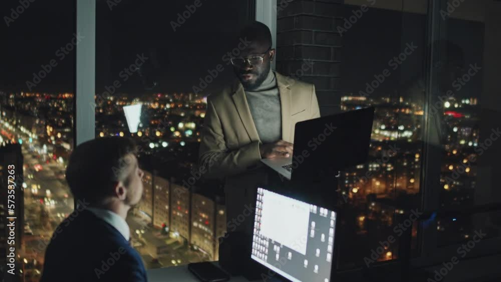Medium shot of Caucasian manager in suit typing on computer late at night, Afro-American colleague with laptop walking into his office to ask for advice, and talking together about work issues Medium shot of Caucasian manager in suit typing on computer late at night, Afro-American colleague with laptop walking into his office to ask for advice, and talking together about work issues