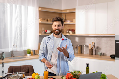 Glad attractive adult caucasian man in casual cooks dinner, juggles tomatoes in modern kitchen interior