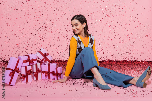full length of joyful woman in trendy clothes looking at pile of gift boxes while sitting near confetti on pink background.