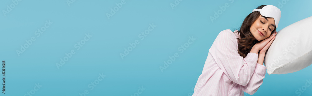 pleased young woman in pajamas and night mask sleeping on white pillow isolated on blue, banner. pleased young woman in pajamas and night mask sleeping on white pillow isolated on blue, banner.