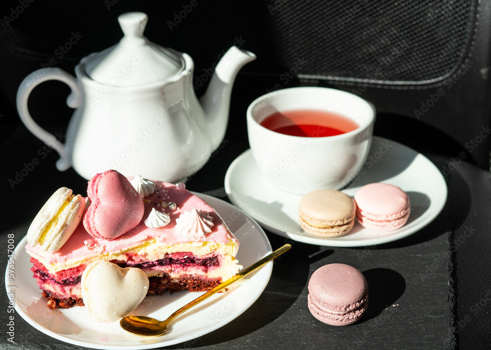 Pink cake with heart shaped macaroons , red tea, white kettle and gold spoon on a black background. Natural light, sunny day