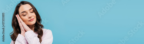 sleepy young woman with closed eyes folding hands while standing in pajamas isolated on blue, banner.