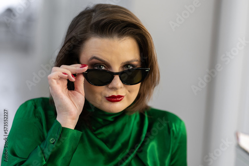 Portrait of attractive beautiful fashionable young woman dressed in the style of 1980s, with bright fancy make up, red nails, glasses, green silk shirt. 80s inspiration and vibes