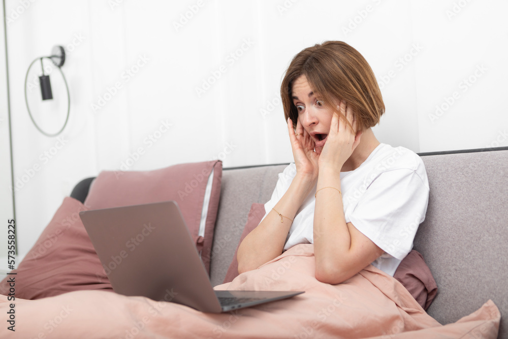 Shocked woman with hands on head working on laptop in the bed. Surprised and upset girl looking at her computer in the bedroom Shocked woman with hands on head working on laptop in the bed. Surprised and upset girl looking at her computer in the bedroom