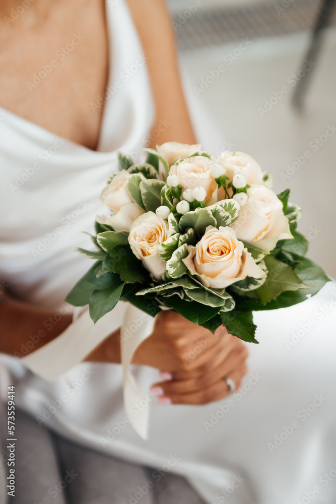 Bride woman holds a wedding bouquet in her hands on the background of white dress. Wedding ceremony. Beautiful beige roses flower and green leaves. Tender sensual close up