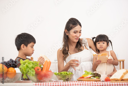 Mom with her two children eating fruits and vegetables. Mother with daughter and toddler son having breakfast at home. Happy lifestyle family. Mother with her children in kitchen cooking together.