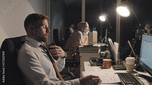 Medium side shot of tired male Caucasian analyst sitting at desk at night in corporate office, studying legal paperwork, loosening tie and rubbing forehead, and female colleague in hijab in background