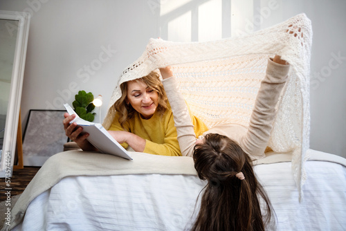 Mature woman liying on bed with happy grandchild girl while reading book and having fun together in cozy bedroom at home. Happy children with grandmother. photo