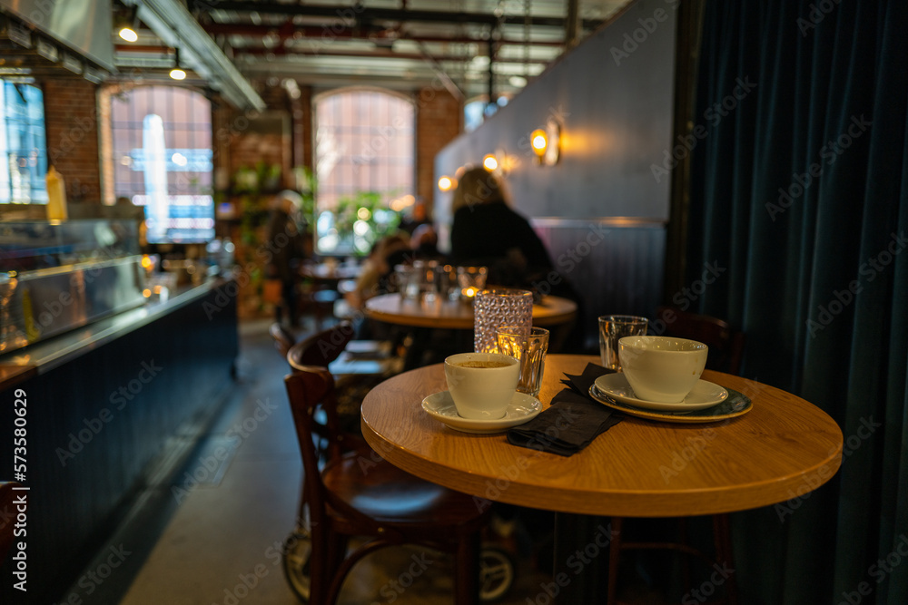 Indoor restaurant setup with empty coffee mugs and glasses on the wooden table with people sitting in the background Indoor restaurant setup with empty coffee mugs and glasses on the wooden table with people sitting in the background