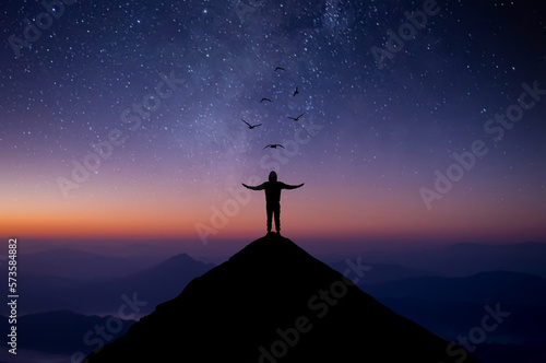 Silhouette of young man standing alone on top of mountain and raise both arms praying and free bird enjoying nature on beautiful night sky, star, milky way background. Demonstrates hope and freedom.