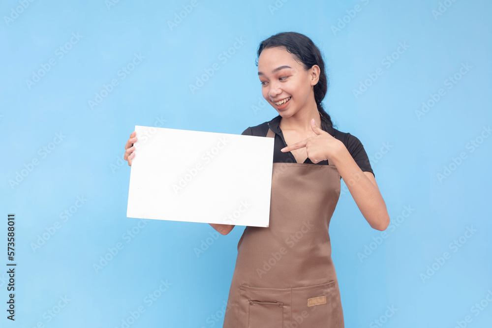 A young asian female waitress or barista points to a blank white signage she is holding. Wearing a brown apron and black shirt welcoming a customer and against a light blue background. A young asian female waitress or barista points to a blank white signage she is holding. Wearing a brown apron and black shirt welcoming a customer and against a light blue background.