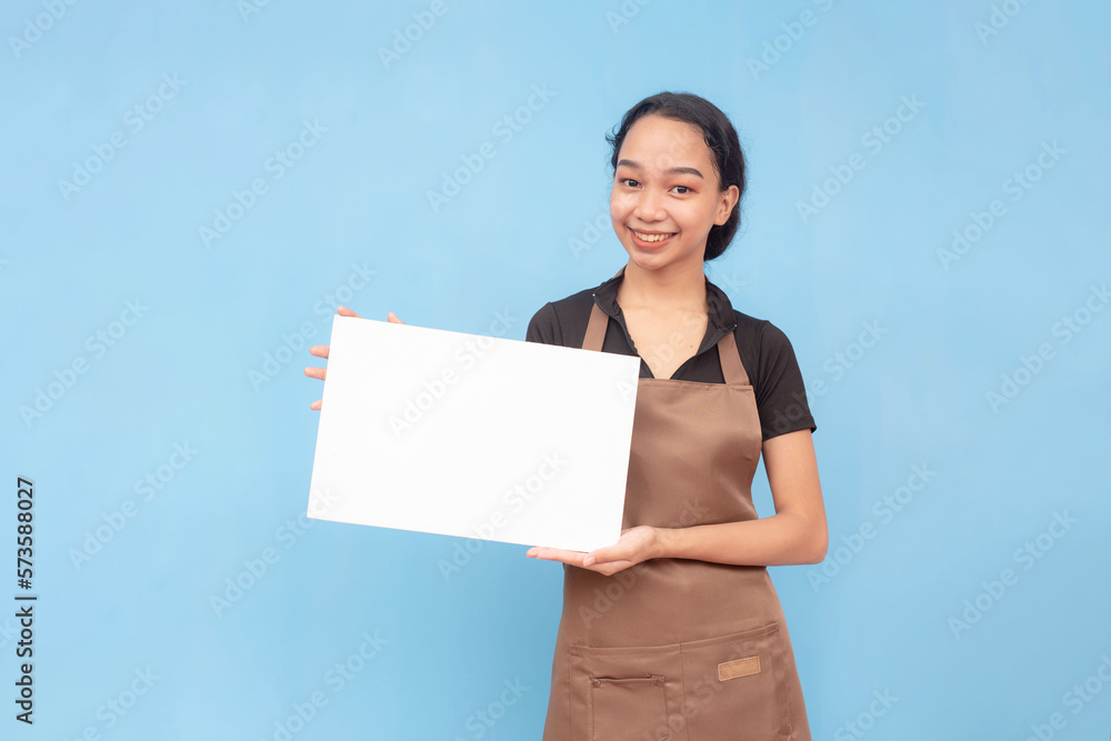 A young asian female waitress or barista holding a blank white signage. Wearing a brown apron and black shirt welcoming a customer and against a light blue background. A young asian female waitress or barista holding a blank white signage. Wearing a brown apron and black shirt welcoming a customer and against a light blue background.