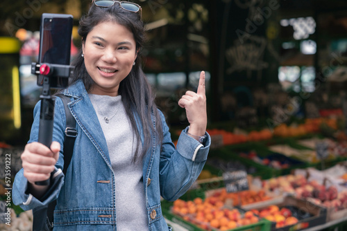 asian female blogger vlogger record vlog streaming video hold phone on selfie stick in open market urban city.Young Asian female tourist woman holding smartphone and recording videos travel concept photo