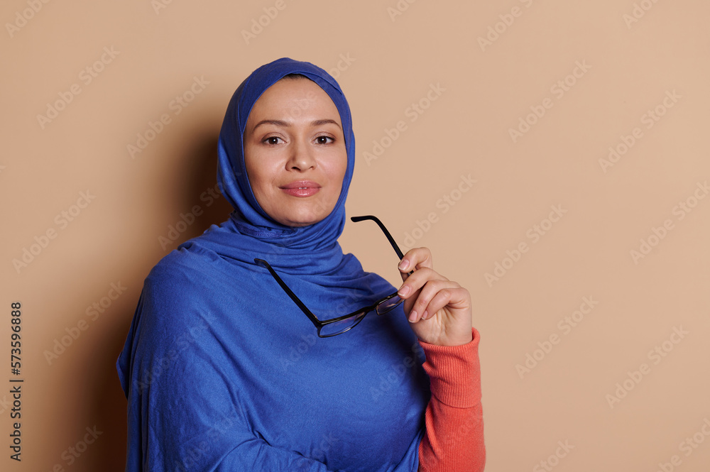 Attractive, confident, modern, stylish Middle-Eastern Muslim woman, business person, teacher, successful entrepreneur in blue hijab, holding spectacles in hand, smiling at camera over beige background Attractive, confident, modern, stylish Middle-Eastern Muslim woman, business person, teacher, successful entrepreneur in blue hijab, holding spectacles in hand, smiling at camera over beige background