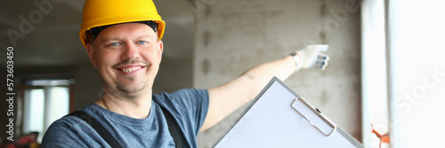 Smiling builder worker in hard hat holding blank sheet of paper on clipboard