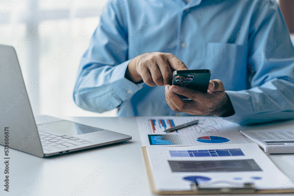 Fototapeta premium Male businessman hand using smartphone to analyze and calculate annual income and expenditure. Financial and technology charts and graphs showing market dynamics and holding smartphone at office