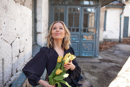 A  beautiful woman dressed in a fashion dress, with a bouquet of  yellow flowers in her hands walking along the old town with ancient buildings of the last century © Вероника Зеленина