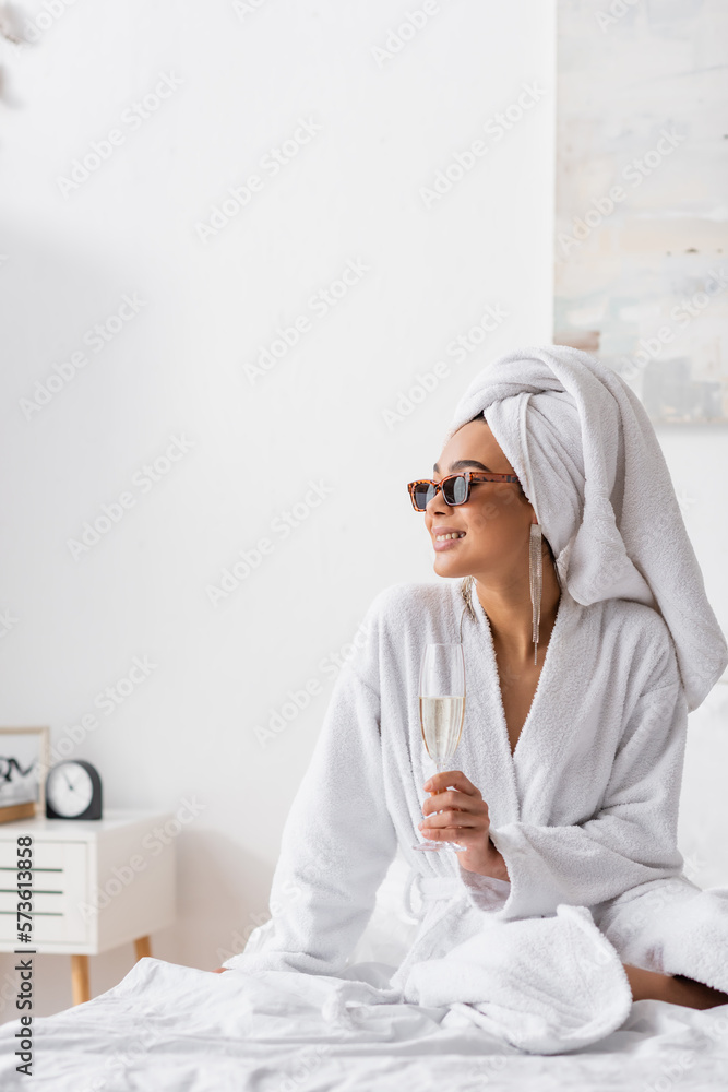 smiling african american woman in terry bathrobe and trendy sunglasses holding champagne and looking away in bedroom. - obrazy, fototapety, plakaty 