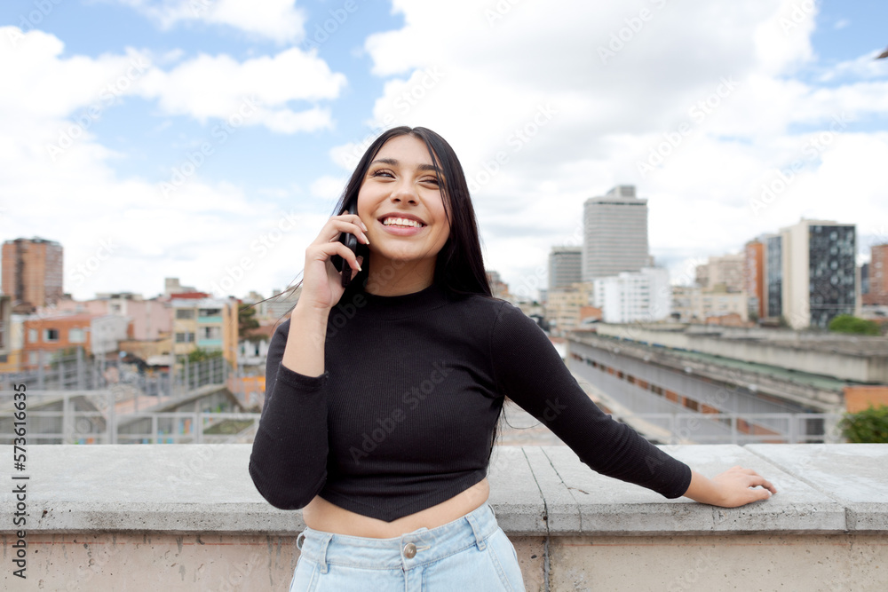 Fototapeta premium Panoramic view of a beautiful smiling woman talking on the phone on a terrace in Bogota with the cityscape in the background, Colombia.