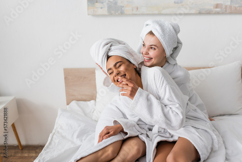 excited african american woman in white bathrobe and towel laughing with closed eyes near happy friend on bed.