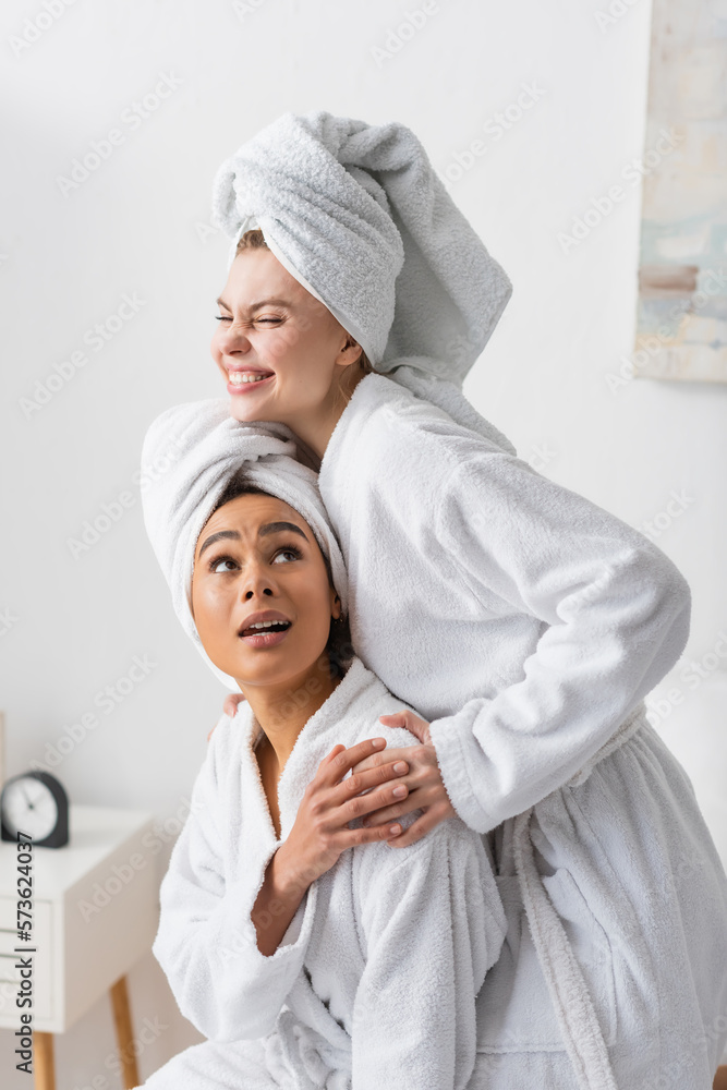 cheerful young woman in white bathrobe and towel embracing african american friend in bedroom. cheerful young woman in white bathrobe and towel embracing african american friend in bedroom.