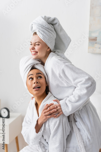 cheerful young woman in white bathrobe and towel embracing african american friend in bedroom.