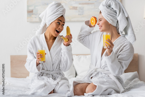 excited multiethnic women with fresh fruits and orange juice having fun while sitting on bed.