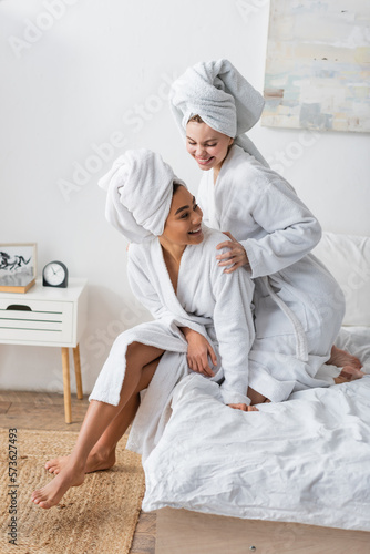 happy woman in white robe and towel embracing african american friend in bedroom.