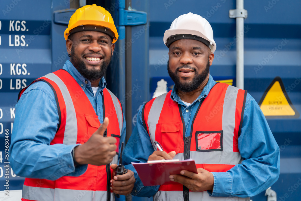 Portrait of Two African Engineer or foreman wears PPE checking container storage with cargo container background at sunset. Logistics global import or export shipping industrial concept.