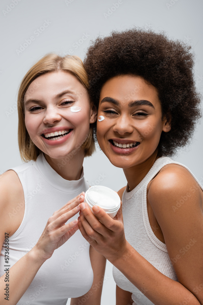 joyful interracial women in white tank tops looking at camera near jar of face cream isolated on grey. joyful interracial women in white tank tops looking at camera near jar of face cream isolated on grey.