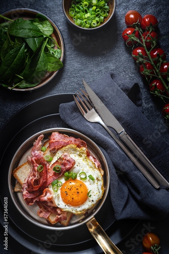 Fototapeta Naklejka Na Ścianę i Meble -  Fried eggs with bacon and green onions in a frying pan. Dark  background. Rustic breackfast. Top view. Rustic served table.