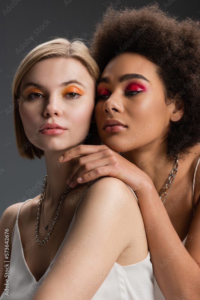 stylish multiethnic models in colorful and glossy eye shadows and white strap dresses posing isolated on grey. stylish multiethnic models in colorful and glossy eye shadows and white strap dresses posing isolated on grey.