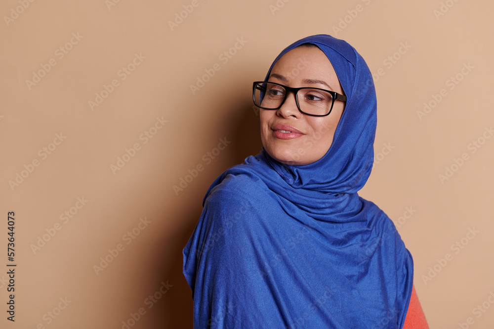 Attractive confident Middle-Eastern Muslim woman in blue hijab, squinting looking to the side through her stylish eyeglasses, isolated on cream background. Vision problems concept. Copy space for ads Attractive confident Middle-Eastern Muslim woman in blue hijab, squinting looking to the side through her stylish eyeglasses, isolated on cream background. Vision problems concept. Copy space for ads