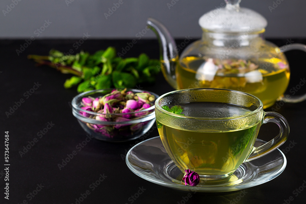cup of tea and teapot. Rose buds tea in a glass teapot on the black table. Teapot with tea made from tea rose petals with green mint. copy space cup of tea and teapot. Rose buds tea in a glass teapot on the black table. Teapot with tea made from tea rose petals with green mint. copy space