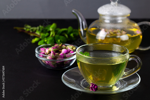 cup of tea and teapot. Rose buds tea in a glass teapot on the black table. Teapot with tea made from tea rose petals with green mint. copy space