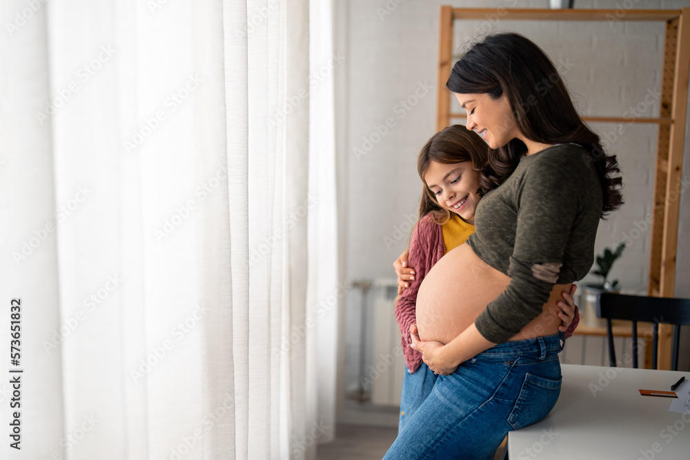 Young smiling little girl hugging pregnant mother, looking at her round belly, thinking about her baby brother or sister while standing near window with white curtains at home during the bright day. Young smiling little girl hugging pregnant mother, looking at her round belly, thinking about her baby brother or sister while standing near window with white curtains at home during the bright day.