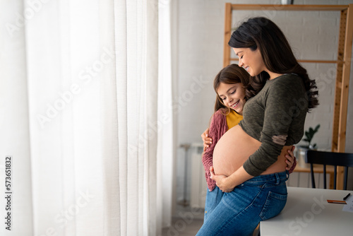 Young smiling little girl hugging pregnant mother, looking at her round belly, thinking about her baby brother or sister while standing near window with white curtains at home during the bright day. © Dorde