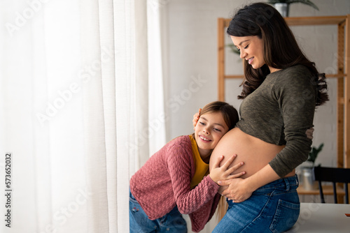 Beautiful pregnant single mother tenderly embracing her sweet little daughter who is leaning head on her pregnant baby bump belly. Curious lovely girl trying to feel the baby inside mother's big belly © Dorde