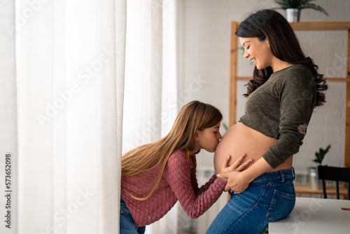 Lovely little girl kissing belly of a pregnant mother. Mother in advanced stage of pregnancy with her adorable girl joyfully bonding at home. Caring little girl giving kisses to baby in mom's tummy.
