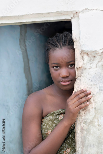 Young Afro girl standing at the door of her house, sad expression, 12 years old, photo